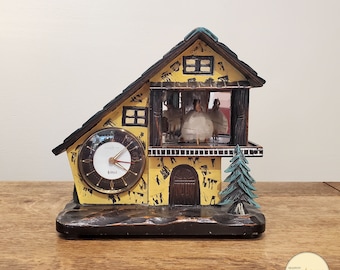 May include: A yellow and brown wooden clock shaped like a house with a clock face and a small pine tree on the side. The clock face has black hands and a white background with black numbers. There are small figures of people inside the house.
