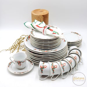 May include: A white Christmas dinner set with gold trim, featuring red and green ribbon and holly designs. The set includes plates, saucers, cups, and a napkin with a matching design. A wooden log and gold decorative beads are also in the image.