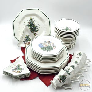 May include: A white Christmas-themed dinnerware set with green trim. Includes octagonal plates, tree-shaped plates, and cups, all decorated with Christmas tree and gift box designs. The set is arranged on a red cloth.