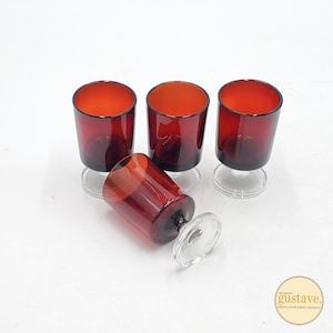 May include: Four vintage red-tinted glass goblets with clear stems. One goblet is lying on its side. The glasses have a rounded shape and are set against a white background. The logo "Maison Gustave" is in the bottom right corner.