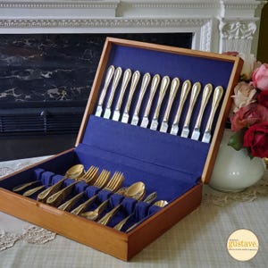 May include: A wooden cutlery case with a blue velvet interior holds a set of gold-colored silverware. The set includes knives, forks, and spoons. The case is open, revealing the arrangement of the utensils. The background includes a fireplace and a vase of flowers.