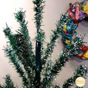 May include: A vintage artificial Christmas tree made of green and silver tinsel. The tree is decorated with a single, large, green and silver tinsel ball at the top.