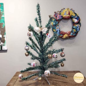 May include: A small artificial Christmas tree decorated with ornaments and a white bow. The tree is on a wooden table with a nativity scene wreath on the wall behind it.