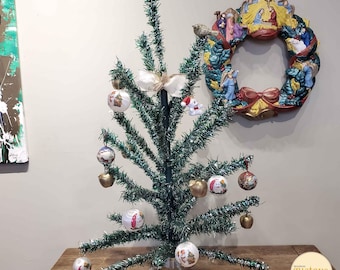 Könnte beinhalten: Ein kleiner künstlicher Weihnachtsbaum, der mit Ornamenten und einer weißen Schleife geschmückt ist. Der Baum steht auf einem Holztisch mit einem Weihnachtskranz mit Krippenszene an der Wand dahinter.