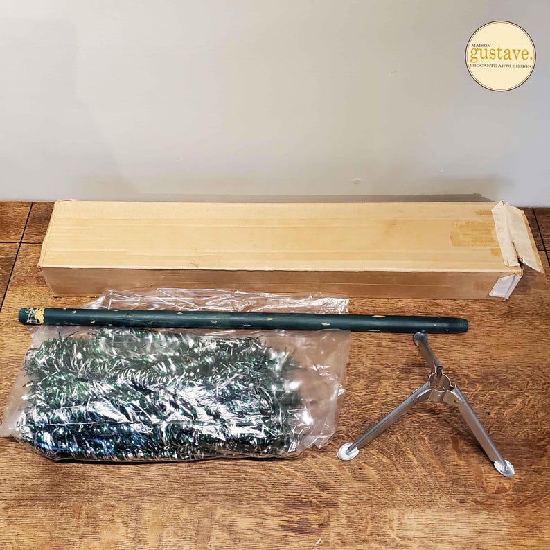 May include: A set of Christmas tree decorations, including a green tinsel garland, a brown cardboard box, a green metal pole, and a silver metal stand.