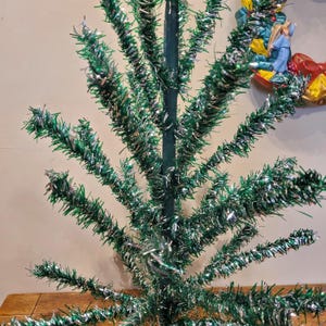 May include: A vintage green and silver tinsel Christmas tree. The tree is made of wire branches covered in tinsel. The branches are curled and twisted to create a realistic tree shape.