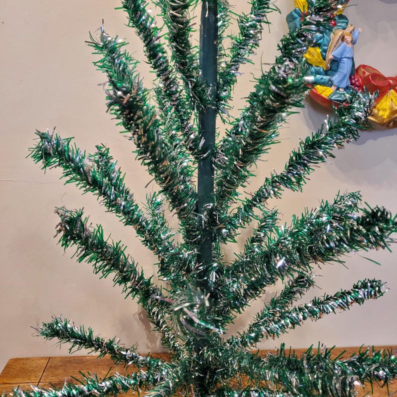 May include: A vintage green and silver tinsel Christmas tree. The tree is made of wire branches covered in tinsel. The branches are curled and twisted to create a realistic tree shape.