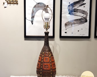 May include: A mid-century modern table lamp with a brown ceramic base featuring a geometric pattern of squares and circles. The base is decorated with gold accents. The lampshade is white and the light bulb is visible.