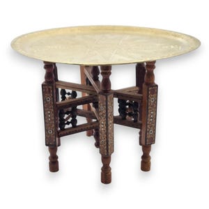 May include: A round, antique brass tray table with a decorative starburst pattern. The table has a dark wood base with inlaid decorative panels and four legs. The table is approximately 61 cm in diameter and 51 cm tall.