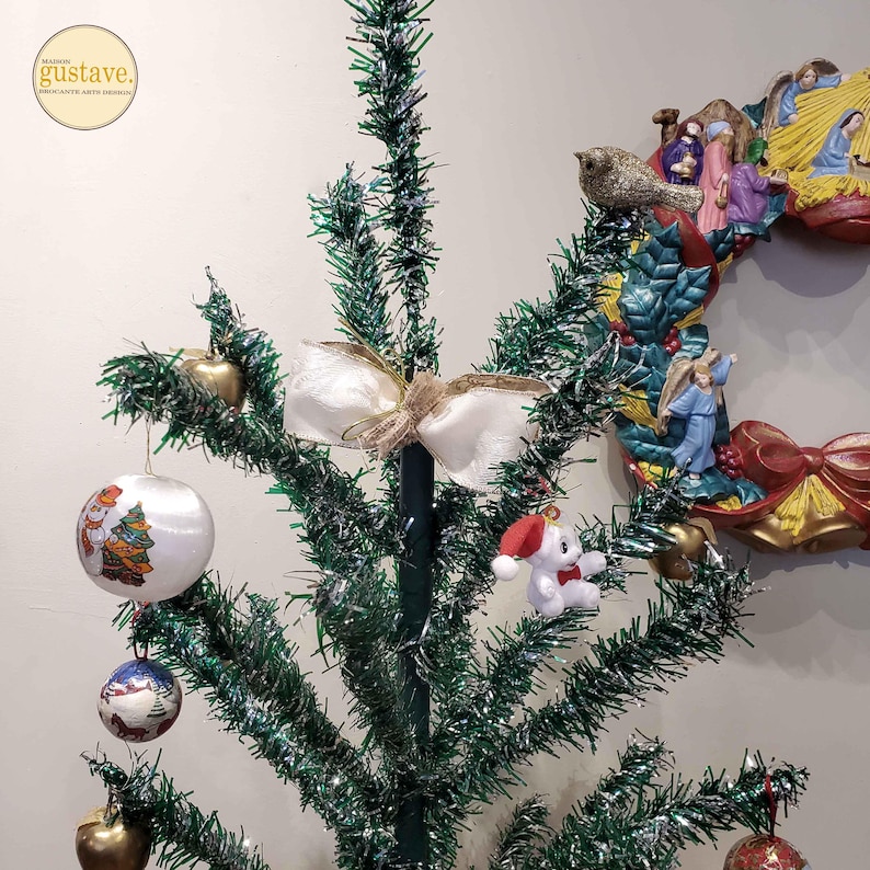 May include: A green tinsel Christmas tree decorated with ornaments, a white bow, and a small stuffed animal wearing a Santa hat.