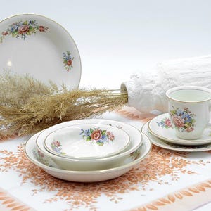 May include: A collection of white porcelain tableware featuring floral patterns and gold rims. The set includes a teacup, saucer, and plates, alongside a white vase with dried plants. The image includes the text "MAISON GUSTAVE. BROCANTE ARTS DESIGN."