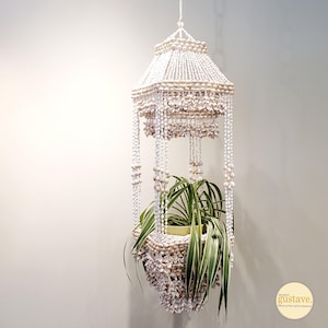 May include: A hanging planter made of white shells, with a pagoda-like top and a lower basket. The planter is suspended by a chain and holds a green plant with long, slender leaves. The shells are beige and white.