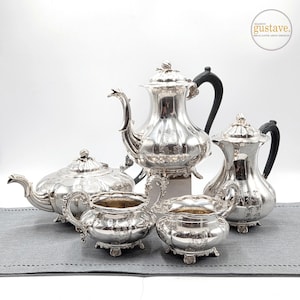 May include: A silver tea set including a teapot, coffee pot, creamer, sugar bowl, and a second teapot. The set has ornate detailing and black handles. The Maison Gustave logo is in the upper right corner.