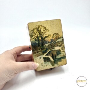 Peut inclure: Une boîte en bois avec une scène peinte d'un pont enneigé et de maisons dans un paysage d'hiver. La boîte a une finition dorée et un aspect légèrement vieilli.