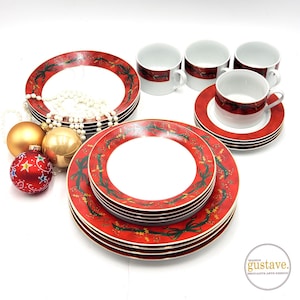 Peut inclure: Un service de table de Noël rouge et blanc. L'ensemble comprend des assiettes, des soucoupes et des tasses avec un motif festif de nœuds verts et de houx. Des boules de Noël dorées et rouges et un collier de perles sont également dans l'image.