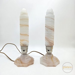 May include: Two vintage table lamps with cylindrical shades and octagonal bases. The shades are white with a textured pattern and streaks of tan. The bases are a light pink with tan veining. Each lamp has a silver-colored socket and a brown electrical cord.