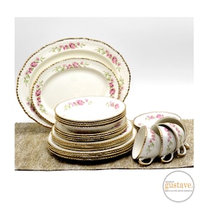 May include: A set of vintage china dinnerware with a floral pattern in shades of pink and green. The plates and bowls have a gold trim and are stacked on a brown surface.