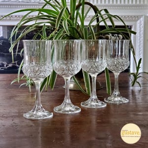 May include: Four clear crystal wine glasses with a detailed pattern on the bowl and a slender stem. The glasses are arranged on a wooden surface, with a green plant and decorative background.