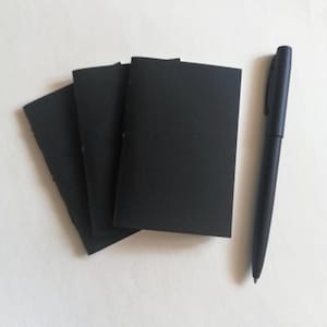 May include: Three small, rectangular notebooks with solid black covers arranged in a fan shape, positioned next to a single black ballpoint pen. The notebooks feature a simple, stitched binding along the spine, and the pen has a matching matte black finish.