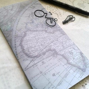 May include: A rectangular notebook with a vintage world map print in shades of gray and white. Resting on the cover are a black ink pen, a black wire paperclip shaped like a bicycle, and a black wire paperclip shaped like a lightbulb.