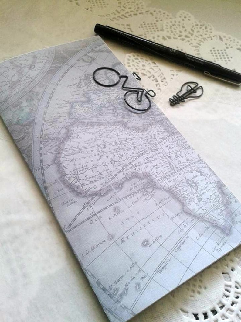 May include: A rectangular notebook with a vintage world map print in shades of gray and white. Resting on the cover are a black ink pen, a black wire paperclip shaped like a bicycle, and a black wire paperclip shaped like a lightbulb.