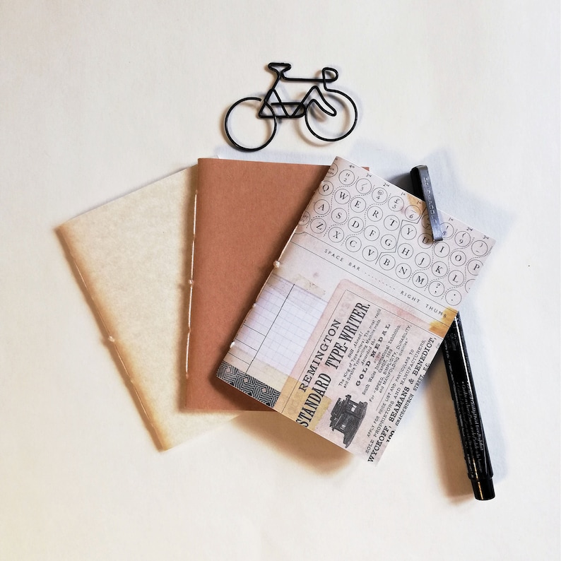 Puede incluir: Un diario marr&oacute;n y crema con un gr&aacute;fico de teclas de m&aacute;quina de escribir y un anuncio vintage de la Remington Standard Type-Writer. El diario est&aacute; encuadernado con hilo y tiene una cubierta de papel marr&oacute;n.