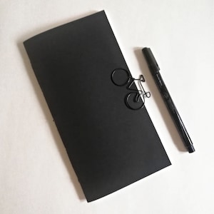 May include: A black notebook with a black bicycle-shaped paperclip and a black pen on a white surface.