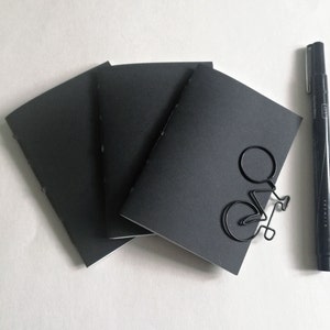 Puede incluir: Tres cuadernos negros con un clip de papel negro con forma de bicicleta en el cuaderno de arriba. Un bol&iacute;grafo negro est&aacute; en el lado derecho de la imagen.