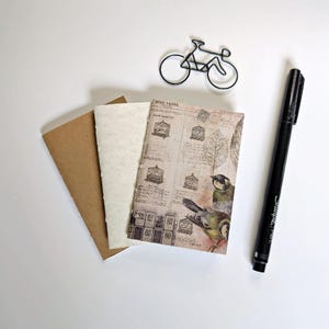 May include: A small notebook with a cover featuring bird cage illustrations, text, and two birds, alongside a plain cream page and a brown cardstock cover. A black wire bicycle-shaped paperclip and a black Sharpie pen are positioned nearby.