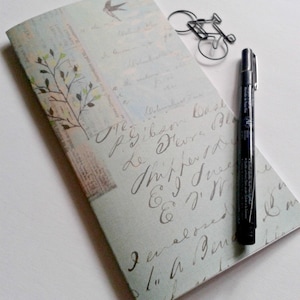 May include: A rectangular notebook with a light blue cover featuring vintage-style cursive script, a small bird illustration, and a tree branch graphic. A black fine-liner pen and a wire bicycle-shaped paperclip rest on the notebook cover.