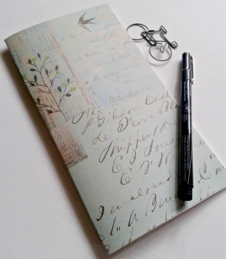 May include: A rectangular notebook with a light blue cover featuring vintage-style cursive script, a small bird illustration, and a tree branch graphic. A black fine-liner pen and a wire bicycle-shaped paperclip rest on the notebook cover.