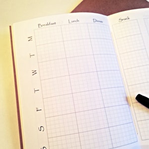 May include: An open planner with white pages featuring a grid layout for meal tracking. The columns are labelled Breakfast, Lunch, Dinner, and Snack. The rows are marked with the letters M, T, W, T, F, S, S. A black pen rests on the right page.