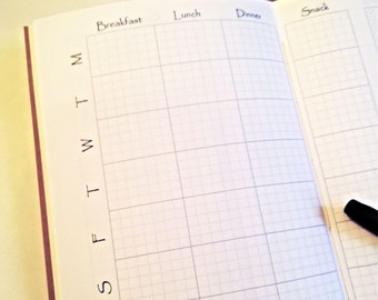 May include: An open planner with white pages featuring a grid layout for meal tracking. The columns are labeled Breakfast, Lunch, Dinner, and Snack. The rows are marked with the letters M, T, W, T, F, S, S. A black pen rests on the right page.