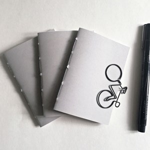Puede incluir: Tres cuadernos grises cosidos con un clip de papel de bicicleta de alambre negro en el cuaderno superior.