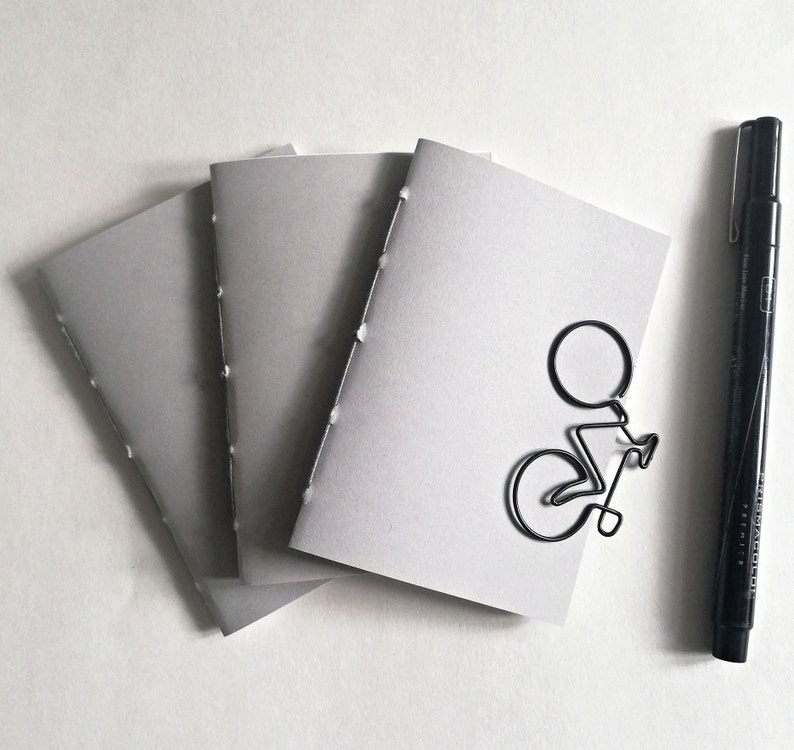 Puede incluir: Tres cuadernos grises cosidos con un clip de papel de bicicleta de alambre negro en el cuaderno superior.