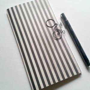 May include: A rectangular notebook featuring a vertical black and white striped pattern. A silver-toned metal paperclip shaped like a bicycle rests on the cover. A black fine-liner pen with a cap is positioned alongside the notebook.