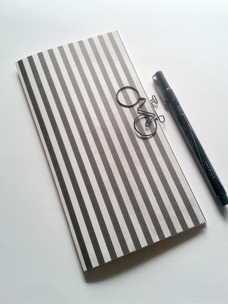 May include: A rectangular notebook featuring a vertical black and white striped pattern. A silver-toned metal paperclip shaped like a bicycle rests on the cover. A black fine-liner pen with a cap is positioned alongside the notebook.