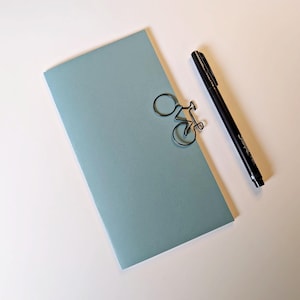 May include: A light blue rectangular envelope with a black metal paper clip shaped like a bicycle attached to the top edge, positioned next to a black felt-tip pen. The items are arranged on a plain surface, showcasing a clean and simple aesthetic.