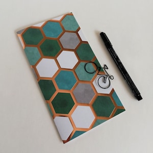 Peut inclure: Une carte rectangulaire avec un motif hexagonal répétitif dans des tons de bleu sarcelle, vert foncé, blanc et gris, séparés par des lignes cuivrées. Un trombone noir en forme de vélo repose sur la carte à côté d'un stylo feutre noir.