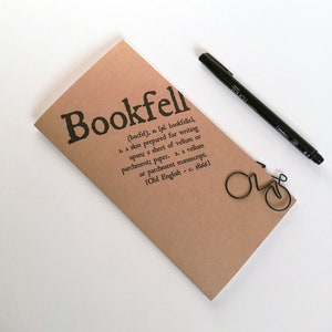 May include: A tan-colored notebook with the word "Bookfell" printed in black, along with a definition. A black pen and a wire bicycle-shaped paperclip are also present. The notebook is a writing accessory.