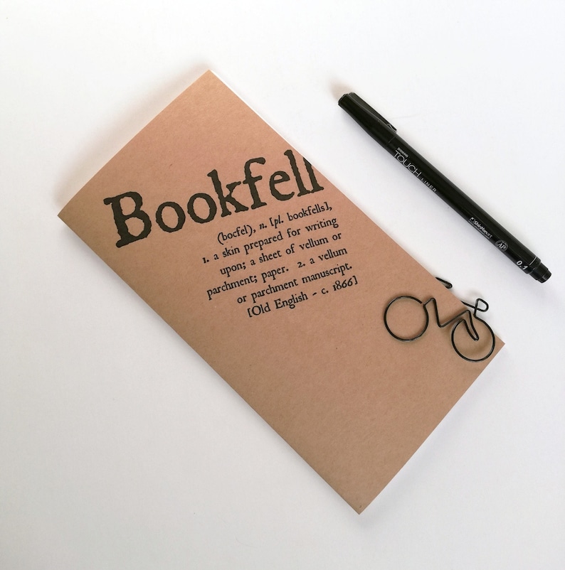 May include: A tan-colored notebook with the word "Bookfell" printed in black, along with a definition. A black pen and a wire bicycle-shaped paperclip are also present. The notebook is a writing accessory.