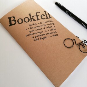 May include: A brown notebook with the word "Bookfell" printed in black, along with a definition. A black pen and a bicycle-shaped paper clip are also visible. The notebook is made of paper.