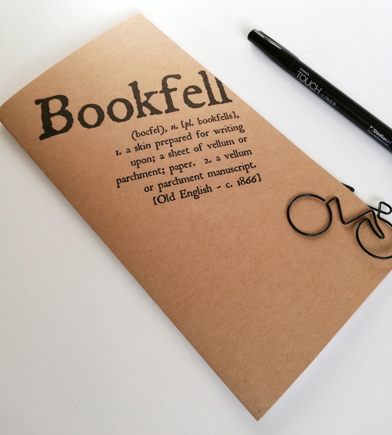 May include: A brown notebook with the word "Bookfell" printed in black, along with a definition. A black pen and a bicycle-shaped paper clip are also visible. The notebook is made of paper.