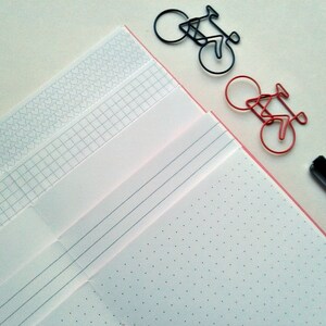 May include: A collection of notebooks with various page styles, including dotted, lined, and grid, alongside bicycle-shaped paper clips in black and red. A black pen rests on the side, suggesting a stationery theme. The notebooks have a pink border.
