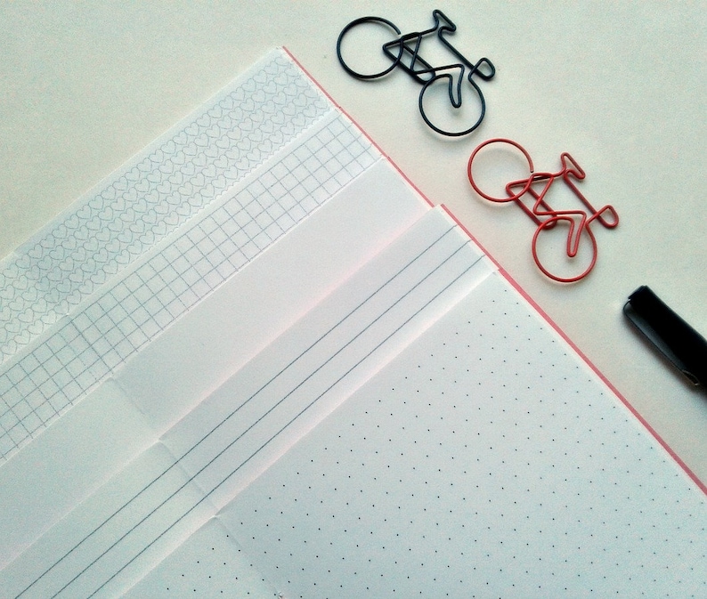 May include: A collection of notebooks with various page styles, including dotted, lined, and grid, alongside bicycle-shaped paper clips in black and red. A black pen rests on the side, suggesting a stationery theme. The notebooks have a pink border.