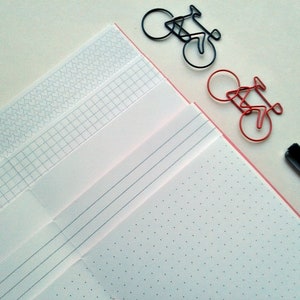 Puede incluir: Un cuaderno blanco con diferentes p&aacute;ginas con patrones. El cuaderno tiene un borde rosa y est&aacute; abierto para revelar una p&aacute;gina con un patr&oacute;n de puntos. Dos clips de papel con forma de bicicletas est&aacute;n en la p&aacute;gina. Uno es rojo y el otro es negro.