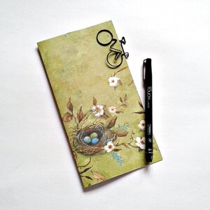 May include: A rectangular notepad with a light green background featuring a painted bird nest with three blue eggs, brown branches, and white flowers. A black bicycle-shaped paperclip is attached to the top, and a black Touch Liner 0.1 pen.