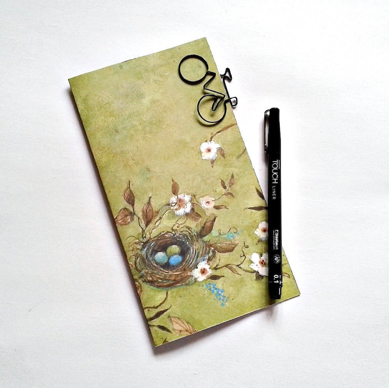 May include: A rectangular notepad with a light green background featuring a painted bird nest with three blue eggs, brown branches, and white flowers. A black bicycle-shaped paperclip is attached to the top, and a black Touch Liner 0.1 pen.