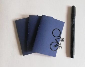 May include: Three small, dark blue notebooks with stitched spines are arranged in a fan shape. A black bicycle-shaped paper clip is attached to the front cover of the top notebook, and a black fine-liner marker is positioned to the right.