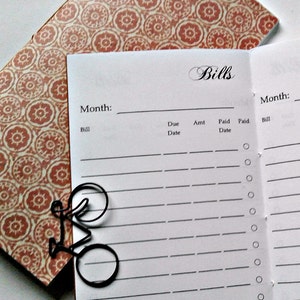 May include: A bill tracker notebook with a white interior page featuring the heading Bills, a Month line, and columns for Bill, Due Date, Amt, Paid Date, and Paid. A black metal bicycle-shaped paperclip rests on the page over a patterned cover.